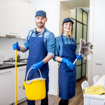 Portrait of a couple as a professional cleaners in uniform standing together with cleaning tools indoors Portrait of a couple as a professional cleaners in uniform standing together with cleaning tools indoors